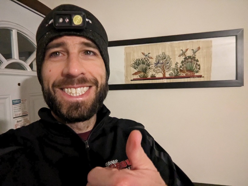 A white man with a dark beard wearing a gray stocking cap, headlamp, and black jacket. He has a wide smile and is giving a thumbs up gesture. Behind him is a framed papyrus painting depicting several Egyptian birds and plants.