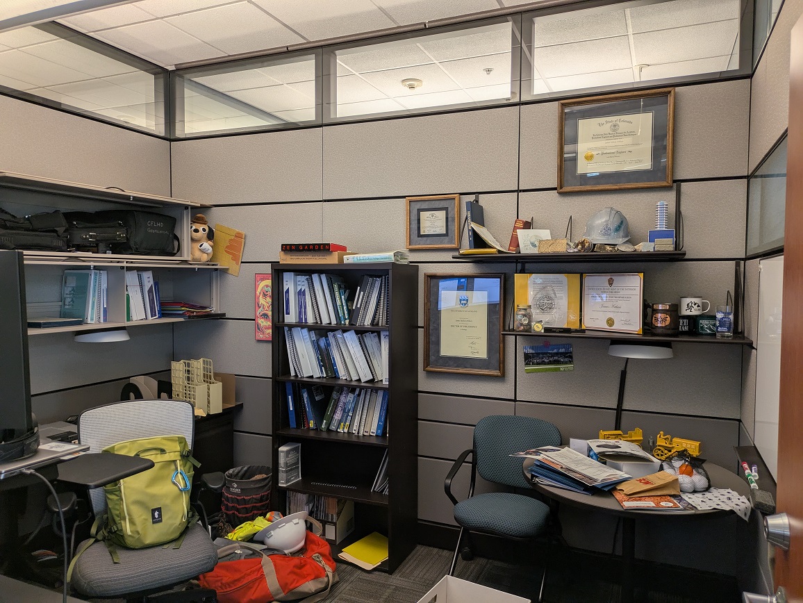A photo of an office with gray walls, several bookshelves full of books and binders, hanging diplomas and awards, and a fair bit of clutter.
